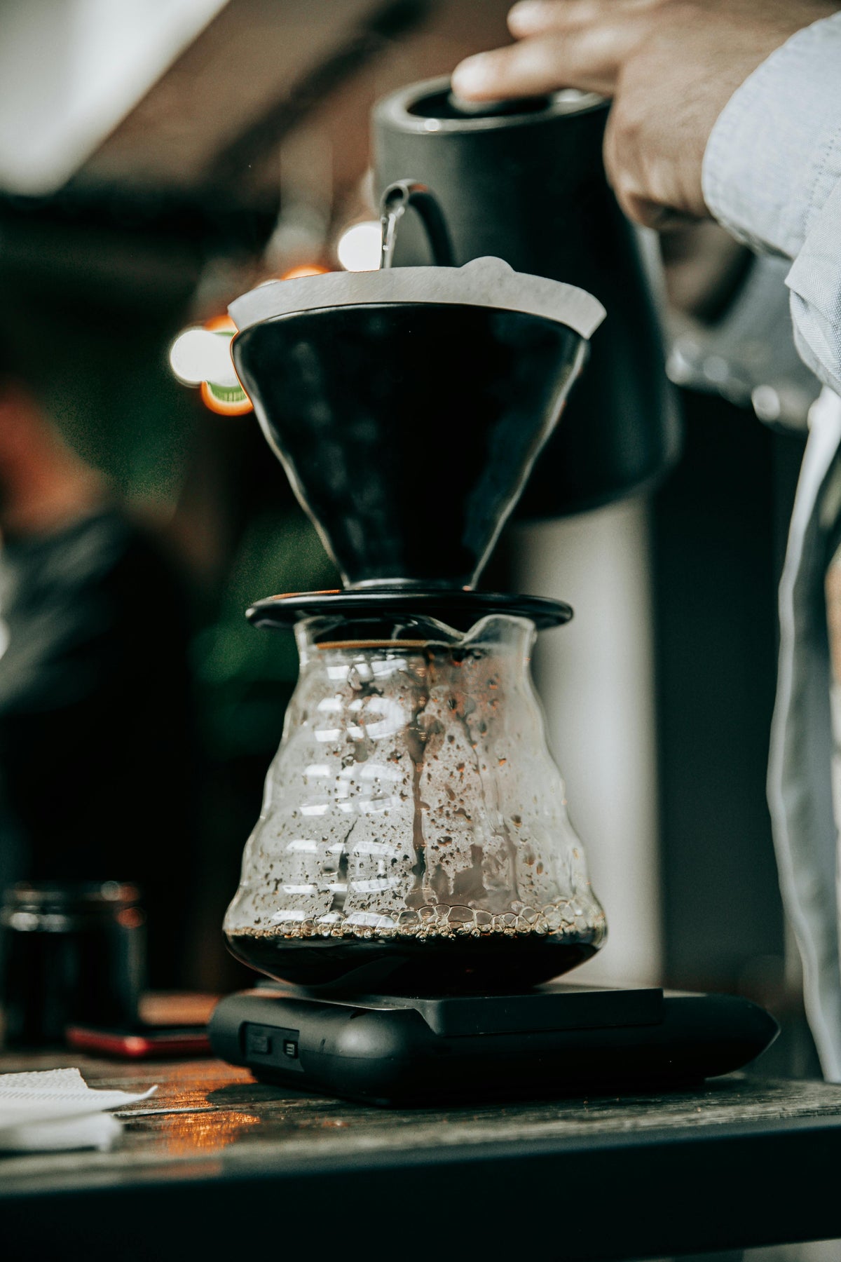 Un individu verse de l'eau avec une bouilloire dans le filtre d'une V60. Le café coule dans la carafe, posée sur une balance.
