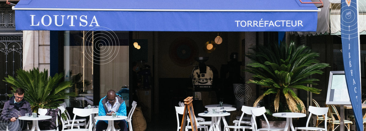 Devanture de la boutique de torréfaction Loutsa, avec terrasse et client assis en extérieur à une table blanche, sous un auvent bleu.
