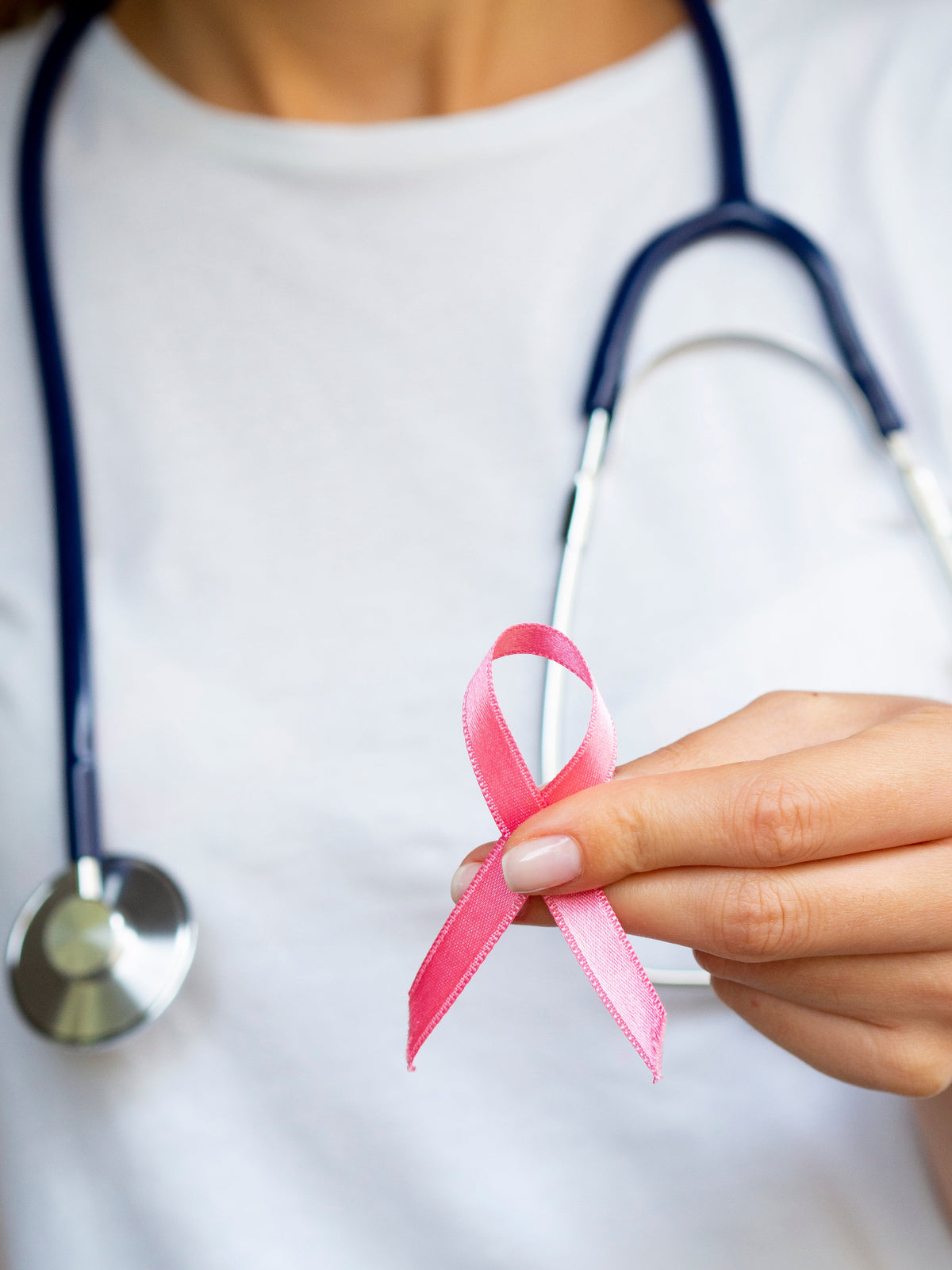 médecin en arrière plan avec un stéthoscope autour du cou, en premier plan sa main qui tient un ruban rose, symbole de la lutte contre les cancers féminins